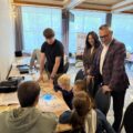 Na zdjęciu dzieci, które skupione są nad kartką papieru, po której poruszają się małe, świecące roboty — Ozoboty. Towarzyszą im uczniowie pełniący rolę opiekunów. Na zdjęciu widoczni są również wójt gminy Janusz Frąckowiak oraz Patrycja Pabich, którzy z zainteresowaniem przyglądają się przebiegowi zajęć.