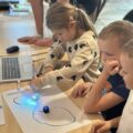 Na zdjęciu widzimy grupkę dzieci skupioną nad kartką papieru, po której poruszają się małe, świecące roboty — Ozoboty. Dzieci rysują trasy, po których poruszają się maszyny. Towarzyszy im starszy opiekun, który obserwuje ich działania.