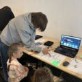 Na fotografii widzimy dzieci skupione nad kartką papieru, po której porusza się mały, świecący robot — Ozobot. Towarzyszy im uczeń pełniący rolę opiekuna, który wskazuje na robota, tłumacząc jego działanie. W tle znajduje się laptop, na którego ekranie widoczne jest programowanie robota.