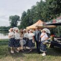 Zdjęcie przedstawia jak nad brzegiem stawku stoi grupa kobiet ubranych w styl retro – w białych bluzkach, granatowych spódnicach w białe grochy, rękawiczkach i kapeluszach z koronkowymi ozdobami. W rękach trzymają wachlarze. Obok nich stoi elegancko ubrany wójt gminy oraz pani sołtys Przemętu. Tło stanowią drzewa i staw.