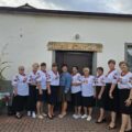 Na zdjęciu grupa kobiet ubranych w białe bluzki z kwiatowymi dodatkami oraz czarne spódnice stoi przed budynkiem, pozując do zdjęcia. Wszystkie mają na sobie czerwone korale. Pomiędzy kobietami stoi sołtys Kluczewa ubrana w elegancki strój. Na budynku wisi drewniane tabliczka z napisem: "Dożynki Kluczewo".