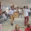 Na zdjęciu widać muzyków występujących na środku sali. Grają m.in. na bębnie i trąbce, a wokół nich uczniowie siedzą w kółku.