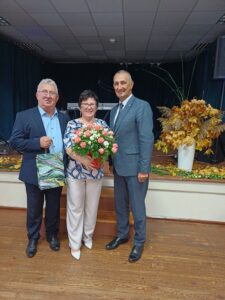Na zdjęciu wójt wraz z dwójką seniorów (kobietą i mężczyzną). Kobieta pozuje z bukietem kwiatów, a mężczyzna z upominkiem.