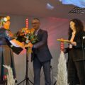 Na zdjęciu wójt wręcza bukiet kwiatów jednej z członkiń KGW. Na zdjęciu także pani Agnieszka Klugiewicz. Wszyscy stoją na scenie.