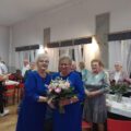 Na zdjęciu dwie kobiety pozują z bukietem kwiatów. W tle inni seniorzy.