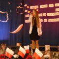 Na zdjęciu widoczna dziewczyna podczas występu na scenie. Scena udekorowana jest w narodowe barwy.