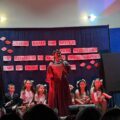 Na zdjęciu widoczna dziewczyna występująca na scenie. W tle inni uczniowie. Scena udekorowana jest w narodowe barwy.
