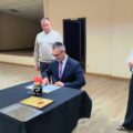 Na zdjęciu widoczny wójt, który siedzi przy stole i podpisuje dokumenty. Na fotografii widoczna też przewodnicząca rady, która stoi obok.