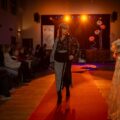 Na zdjęciu widoczna jest kobieta idąca wzdłuż czerwonego dywanu na wybiegu. Ubrana jest w elegancki strój. Na fotografii widać też widownię siedzącą na krzesłach.