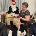 Na zdjęciu widoczni są kobieta oraz mężczyzna przebrany za Świętego Mikołaja, którzy rozdają seniorom świąteczne upominki.