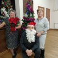 Na fotografii dwie kobiety oraz mężczyzna, który ma ubraną czapkę mikołaja i założoną sztuczną, białą brodę. W tle dwie choinki ozdobione bombkami.