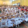 Na zdjęciu widoczne dzieci i osoby dorosłe siedzące na widowni w sali gimnastycznej w szkole. Dzieci przebrane są w stroje związane ze świętami.