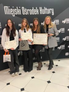 Na zdjęciu dwie uczennice wraz z nauczycielkami. Uczennice pozują z certyfikatami za działalność charytatywną, a nauczycielki z certyfikatem przyznającym szkole tytuł dla najbardziej aktywnej wolontariacko szkoły. W tle ścianka.