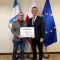 Na fotografii wójt wraz z sołtysem Barchlina pozują z czekiem. W tle widoczne są flagi: Polski, Unii Europejskiej oraz z herbem gminy Przemęt.