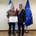 Na fotografii wójt wraz z sołtysem Moch pozują z czekiem. W tle widoczne są flagi: Polski, Unii Europejskiej oraz z herbem gminy Przemęt.