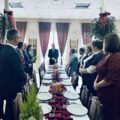 Na fotografii widoczne osoby będące na wigilii. Wszyscy stoją przy dużym stole i słuchają przemowy prezesa konwalii.