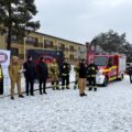 Na zdjęciu widoczni są strażacy, policjanci, przedstawicielki sanepidu oraz wójt. Komendant straży przemawia podczas spotkania.