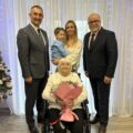 Grupowe zdjęcie, na którym znajdują się wójt, kierownik USC oraz jubilatka wraz z bliskimi. Jubilatka pozuje z bukietem kwiatów.