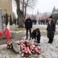 Na fotografii widoczna jest osoba zapalająca znicz przy pomniku, obok którego złożono biało-czerwone wiązanki. W tle stoją uczestnicy uroczystości.