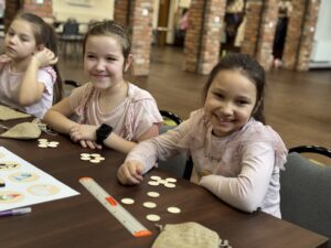 Dwie dziewczynki siedzą przy stole i pozują do zdjęcia. Na stole przedmioty do tworzenia gry "kółko i krzyżyk".